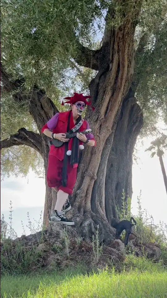 Ryan in red jester motley and skull paint, balanced one-footed on the roots of an old olive tree, playing a ukulele