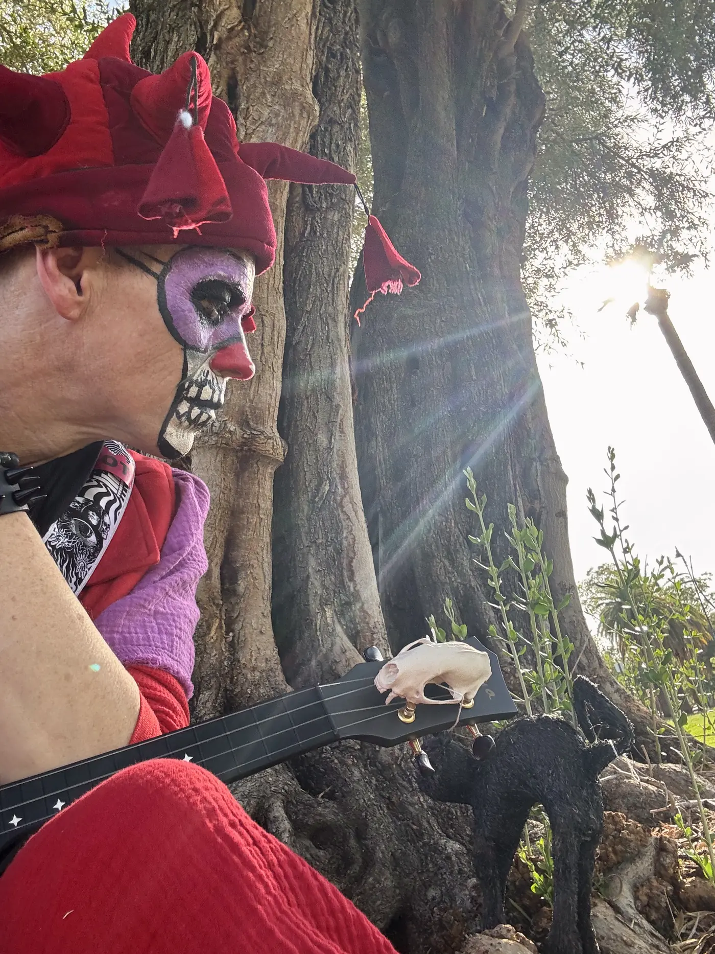 Ryan in jester hat and skull paint, in profile beneath an olive tree, sun flaring through the branches