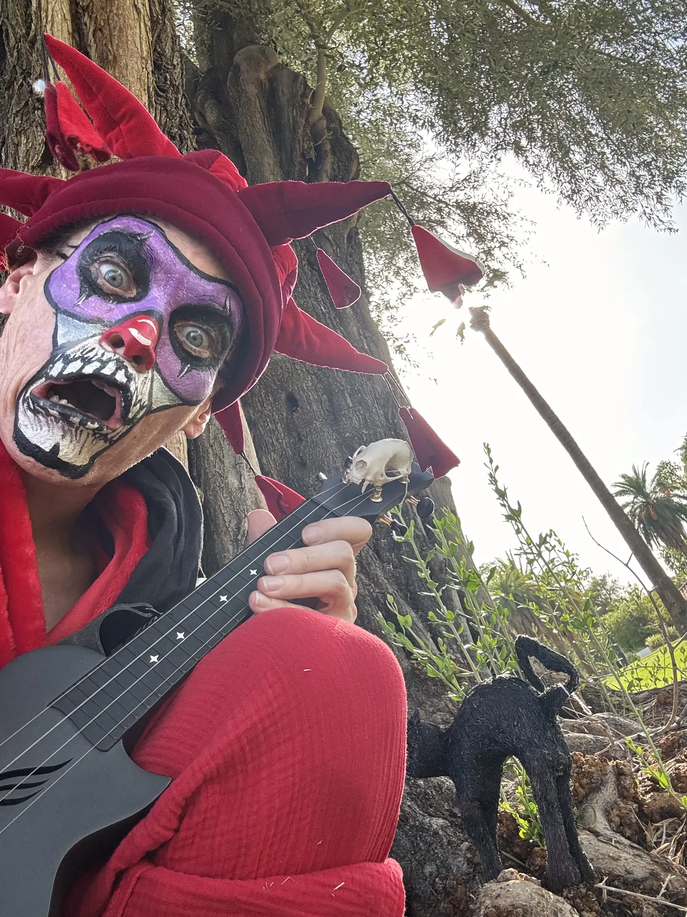 Ryan close to camera in jester hat and skull paint, mouth wide, a ukulele across his knee