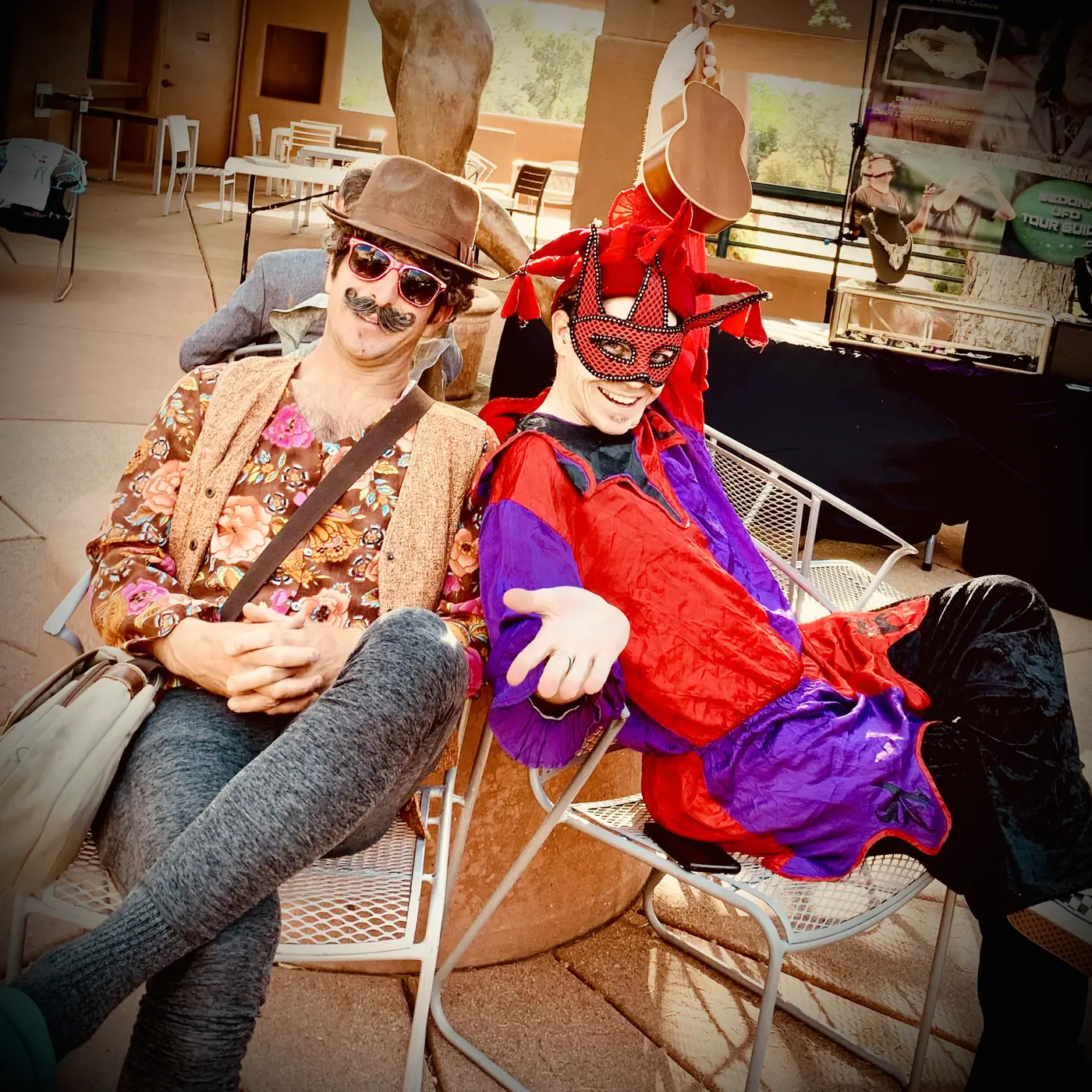Ryan in red and purple motley and a lace mask, sprawled in a patio chair beside a friend in a fedora and false moustache